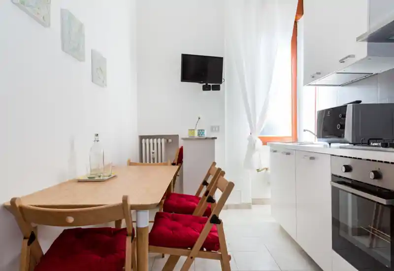 Cuisine compacte et moderne avec placards blancs, four encastré et table en bois clair avec coussins rouges. La lumière de la fenêtre et le style soigné la rendent idéale pour les locations de courte durée.