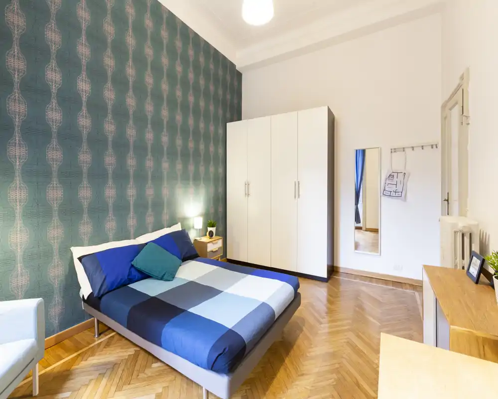 Bright, stylish bedroom with a double bed dressed in blue checked bedding, patterned teal accent wall, wardrobe and wooden parquet flooring — a cozy, well-lit space ideal for relaxing.