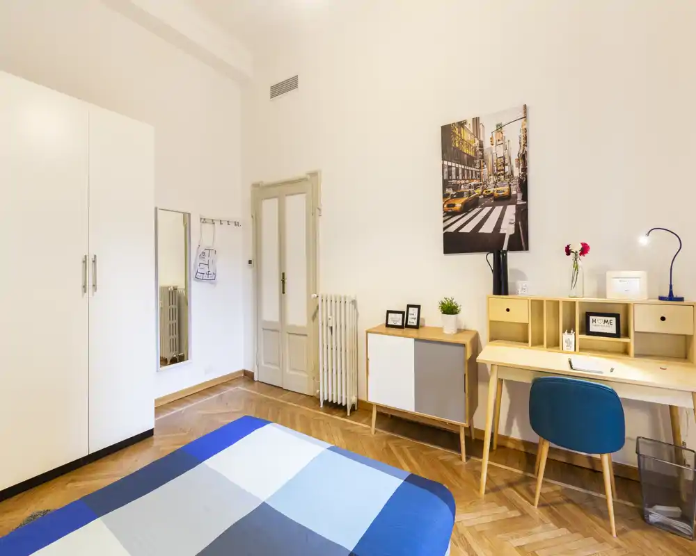 Bright, modern bedroom with a blue-check bedspread, wooden parquet floor, wardrobe and a functional study corner with desk and chair — ideal for a comfortable stay.