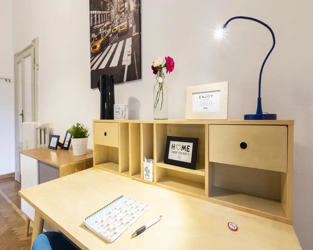 A bright, well-styled living/work corner with a wooden desk, shelving cubbies, decorative prints and a flexible desk lamp — ideal for home office or reading.