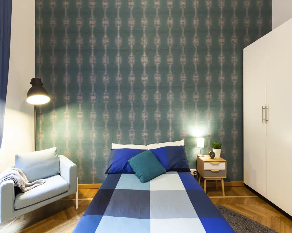 Bright, stylish bedroom with a double bed dressed in blue linens, a bedside table and lamp, wardrobe and a cozy armchair — ideal for a comfortable stay.