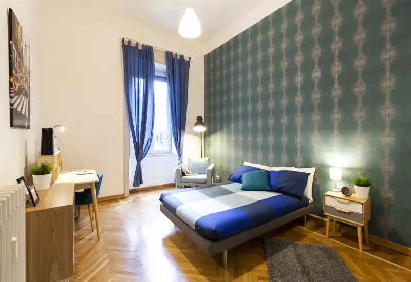 Bright, stylish bedroom with patterned accent wall, double bed dressed in blue linens, bedside table with lamp and a small desk by the window — ideal for a comfortable stay.