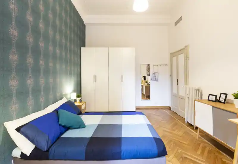 Bright, modern bedroom with a double bed dressed in blue linens, decorative pillows, patterned accent wall and a white wardrobe providing ample storage.