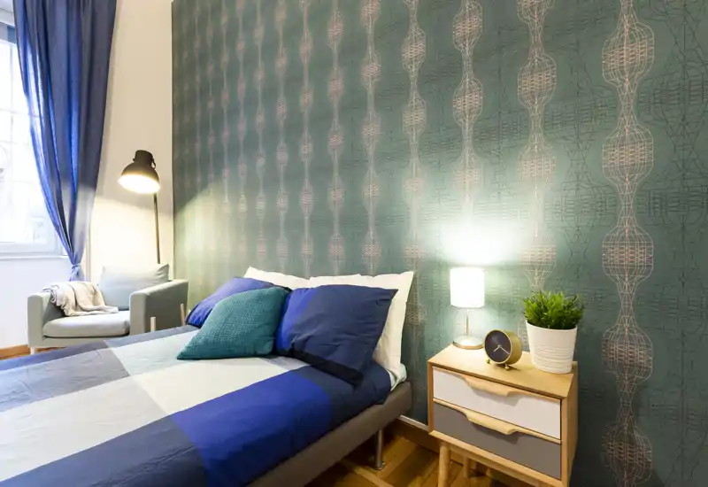 Bright, styled bedroom with a double bed against patterned teal wallpaper, bedside table with lamp and plant, and a cozy armchair by the window.