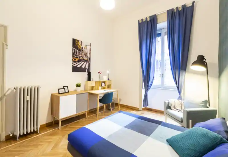 Bright, stylish bedroom with a double bed dressed in blue geometric bedding, a cozy armchair by a standing lamp, and a work desk under a framed photo. Large window with blue curtains provides natural light and warm hardwood floors complete the inviting atmosphere.