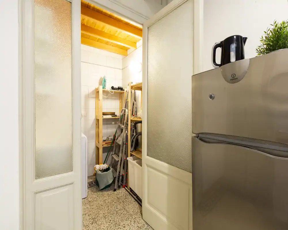 Small kitchen/utility corner with a visible fridge in the foreground and a pantry/storage area with shelves, ladder and cleaning tools behind the glass doors.