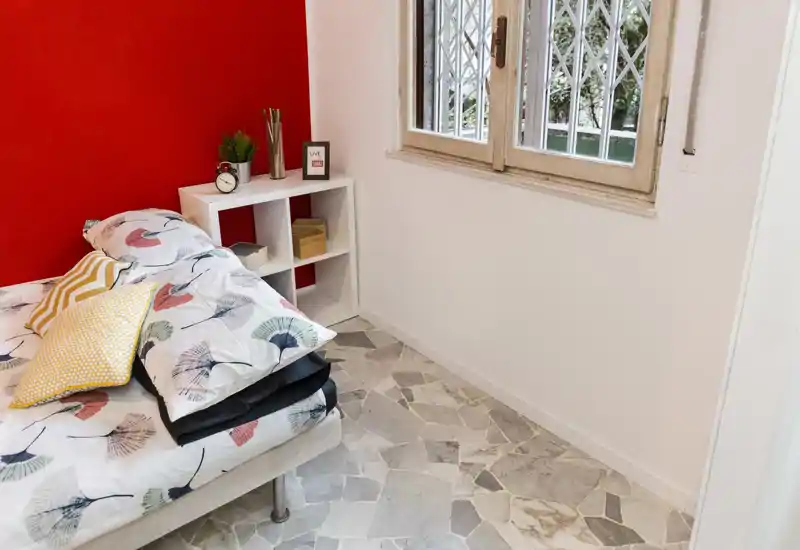 Dormitorio luminoso y ordenado con edredón estampado y cojines decorativos, una pequeña estantería cúbica como mesita de noche y luz natural desde la ventana. La pared roja añade un toque llamativo al espacio sencillo.