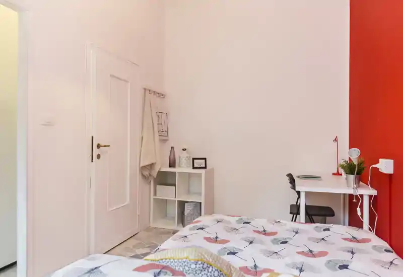 Rincón de dormitorio acogedor y bien iluminado con ropa de cama estampada, pequeña estantería blanca y escritorio compacto junto a una pared roja — ideal para una persona o estudiante.