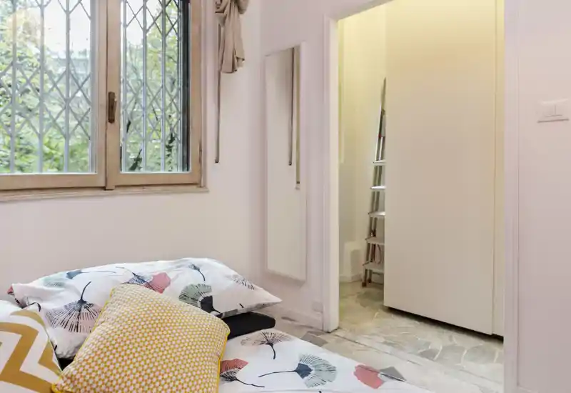 Bright, cozy bedroom corner with patterned bedding and decorative pillows near a window that brings natural light; a doorway leads to an adjoining area.