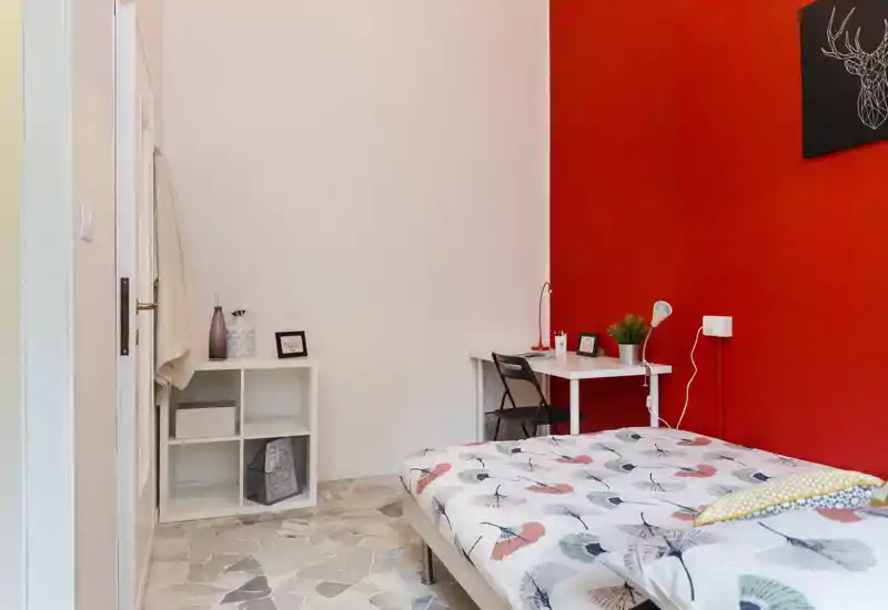 Dormitorio luminoso y bien decorado con cama doble estampada, pequeño escritorio y estantería frente a una pared roja intensa — ideal para una o dos personas.