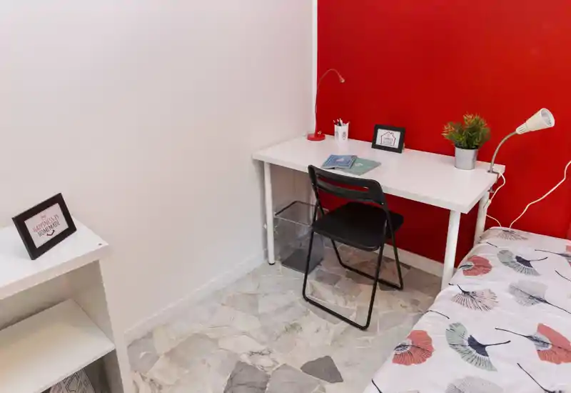 Compact bedroom corner with a single bed partially visible, a white desk with lamp and chair, and simple decorative details like a plant and framed photos. Bright, tidy and suitable for a student or short-term stay.