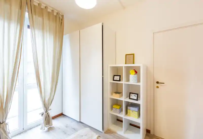 Coin de chambre lumineux avec lumière naturelle d'une grande fenêtre, une armoire blanche et une étagère ouverte avec boîtes et cadres décoratifs ; style moderne et épuré.