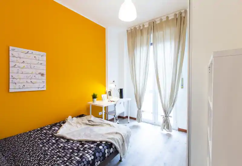 Chambre moderne et lumineuse avec un mur orange, un lit double confortable et un petit bureau près de la fenêtre laissant entrer beaucoup de lumière naturelle.