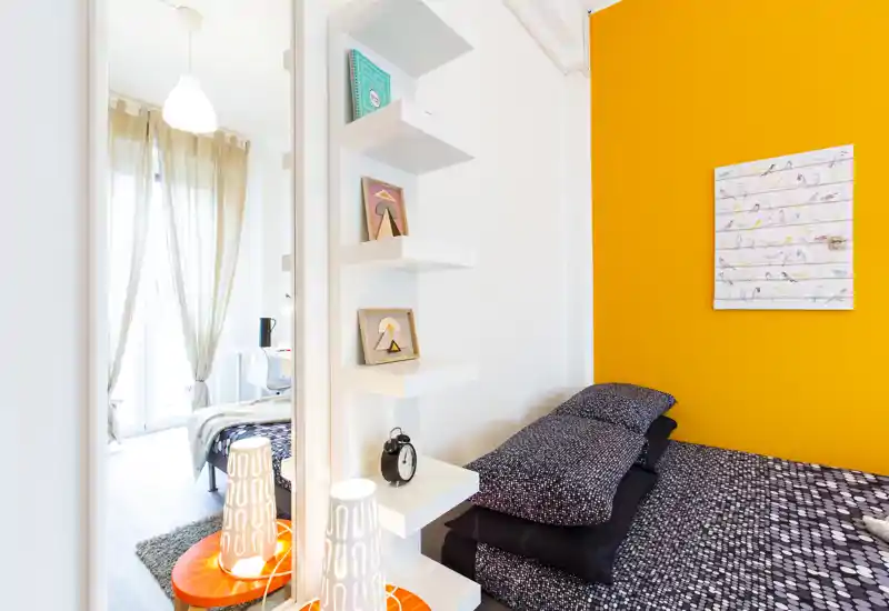 Chambre moderne et lumineuse avec lit à motif, oreillers empilés et un mur orange vif; la lumière naturelle filtre à travers des voilages.