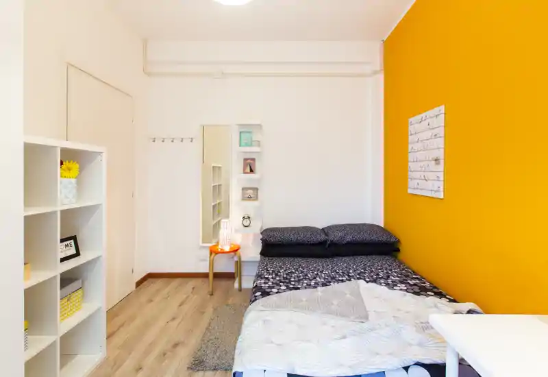 Chambre lumineuse et soignée avec un lit double contre un mur orange, coussins décoratifs et étagères simples — idéale pour un séjour chaleureux.