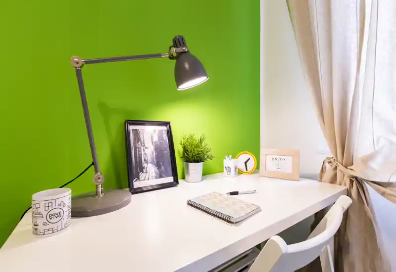 Bright, modern desk corner with a white table, adjustable desk lamp, small potted plant and framed photo against a vivid green wall — ideal for a compact workspace.