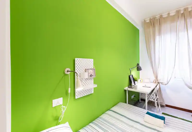 Camera da letto luminosa e moderna con parete verde acceso, letto ben rifatto e una piccola scrivania vicino alla finestra — ideale per uno studente o un viaggiatore singolo.