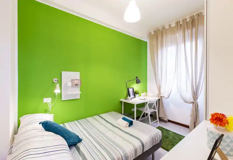 Camera da letto luminosa e moderna con parete verde acceso, letto matrimoniale ben rifatto e una scrivania compatta vicino alla finestra — ideale per studente o professionista.