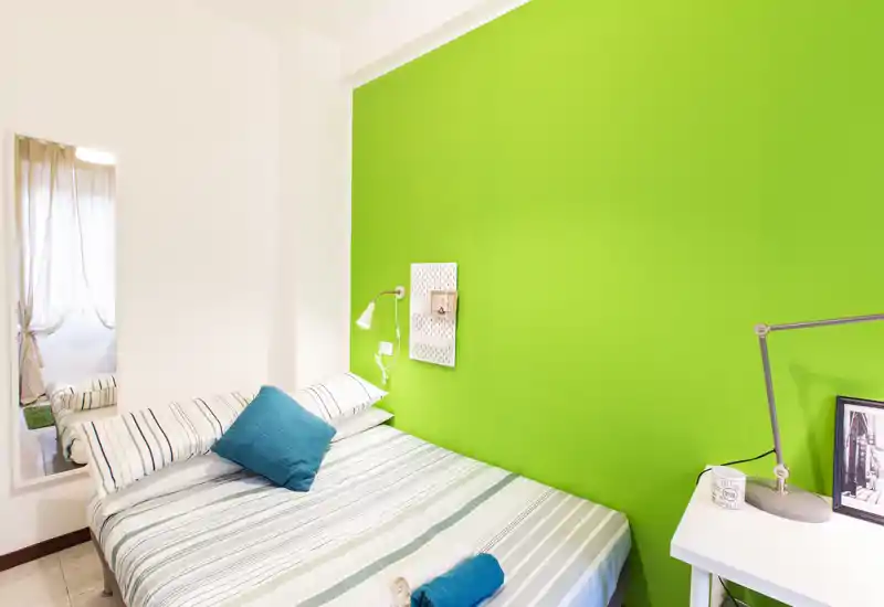 Camera da letto moderna e luminosa con letto matrimoniale a righe contro una vivace parete verde, cuscini accoglienti e un piccolo comodino con lampada — ideale per una coppia o single.
