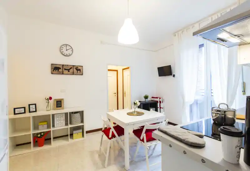 Bright, tidy open-plan dining area with a white table and four chairs with red cushions, integrated with a small kitchen counter and large window providing natural light.