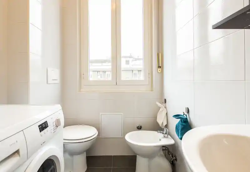 Bright, modern small bathroom with a toilet, bidet, sink and a washing machine under natural window light.