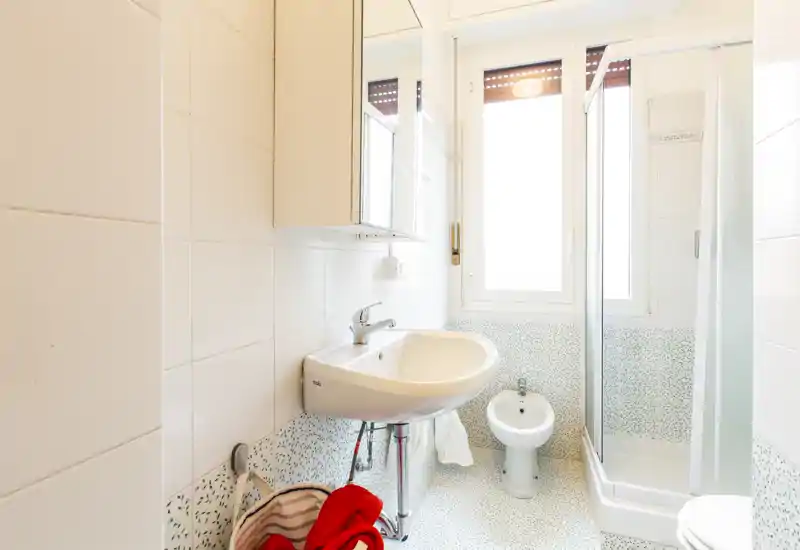 Bright, clean bathroom with a wall-mounted sink, bidet and a corner shower cubicle. Large window provides natural light and white tiles make the space feel airy.