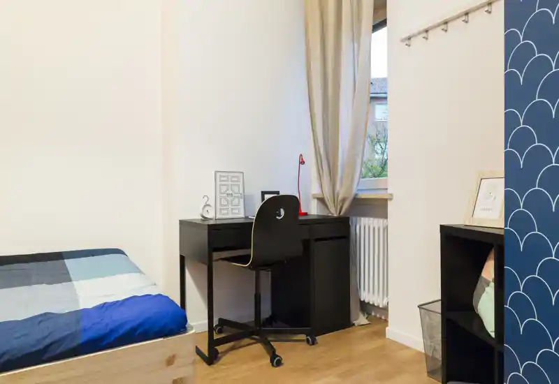 Camera da letto piccola e ordinata con letto singolo e scrivania nera vicino alla finestra. Luce naturale, pavimento in legno e arredo moderno e essenziale, ideale per studente o persona singola.
