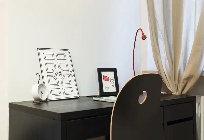 Angolo studio compatto con scrivania in legno scuro, sedia, lampada da lavoro e stampe decorative; luce naturale morbida dalla finestra crea un ambiente tranquillo.