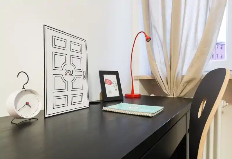 Area di lavoro luminosa e ordinata con scrivania nera, lampada da tavolo rossa e dettagli decorativi accanto a una finestra con tenda.