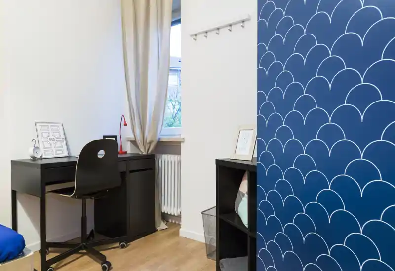 Angolo soggiorno/studio ordinato e luminoso con una scrivania nera compatta, sedia su ruote e scaffalatura decorativa davanti a una parete blu con motivo — ideale per smart working o letture leggere.