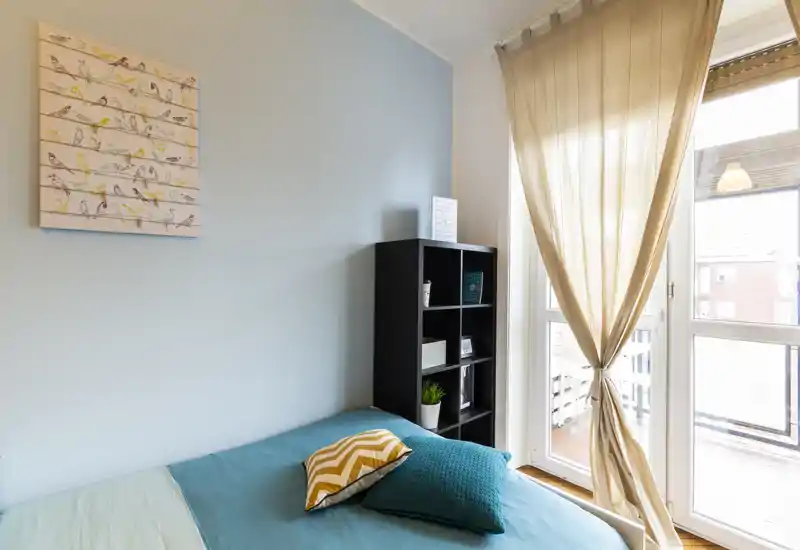 Dormitorio luminoso y ordenado con colcha turquesa, cojines decorativos y una gran ventana que aporta luz natural. Una estantería negra y cortinas suaves completan el estilo contemporáneo y relajante.
