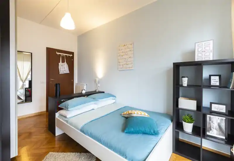 Dormitorio moderno y luminoso con cama doble arreglada en tonos turquesa, lámpara de noche y estantería con objetos decorativos.