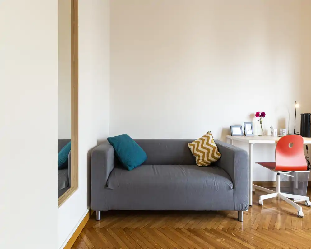 Zona giorno luminosa e moderna con un divano grigio compatto, cuscini colorati e un piccolo angolo scrivania — ideale per un soggiorno accogliente o per lavorare da remoto.