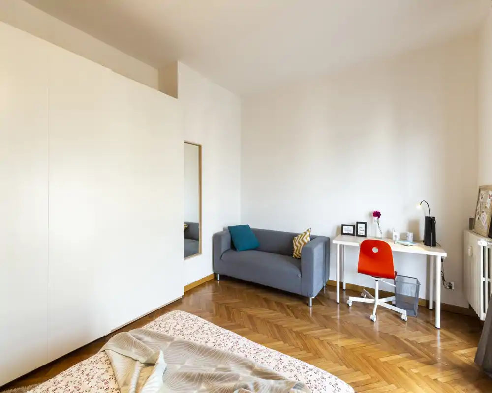 Camera da letto luminosa e ordinata con il letto in primo piano, un divano grigio compatto e una scrivania con sedia arancione — perfetta per una persona o uno studente.