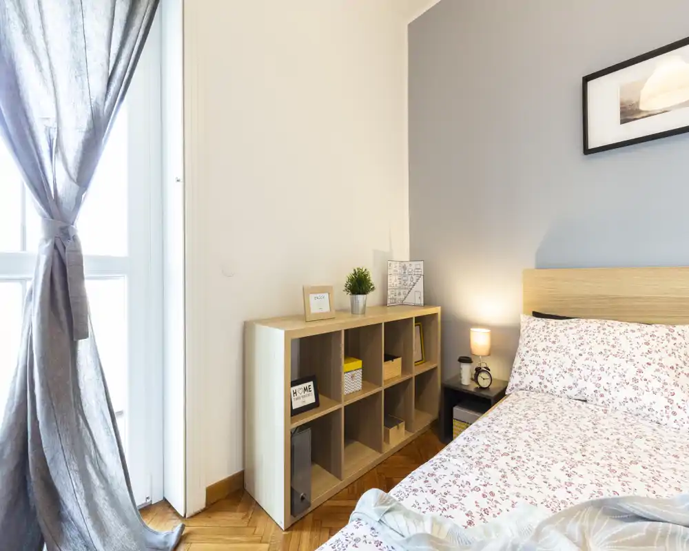 Camera da letto luminosa e accogliente con copriletto a fantasia, comodino con lampada e luce naturale proveniente da una finestra alta con tende.
