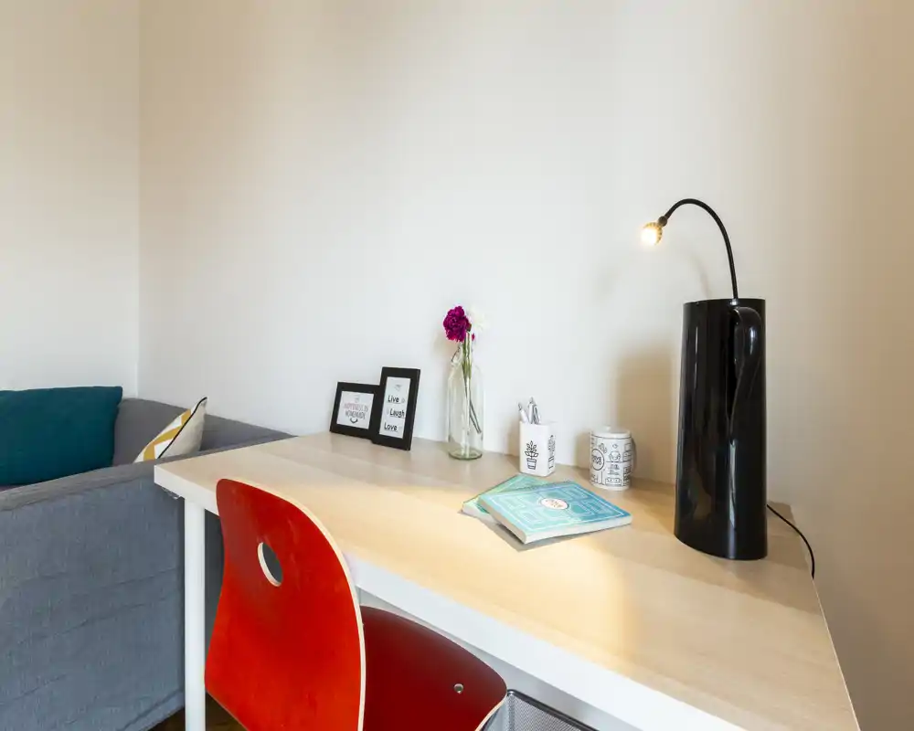 Angolo soggiorno luminoso e ordinato con una scrivania in legno, sedia rossa e divano grigio. Dettagli decorativi come cornici e un vaso con fiori rendono l'ambiente accogliente.