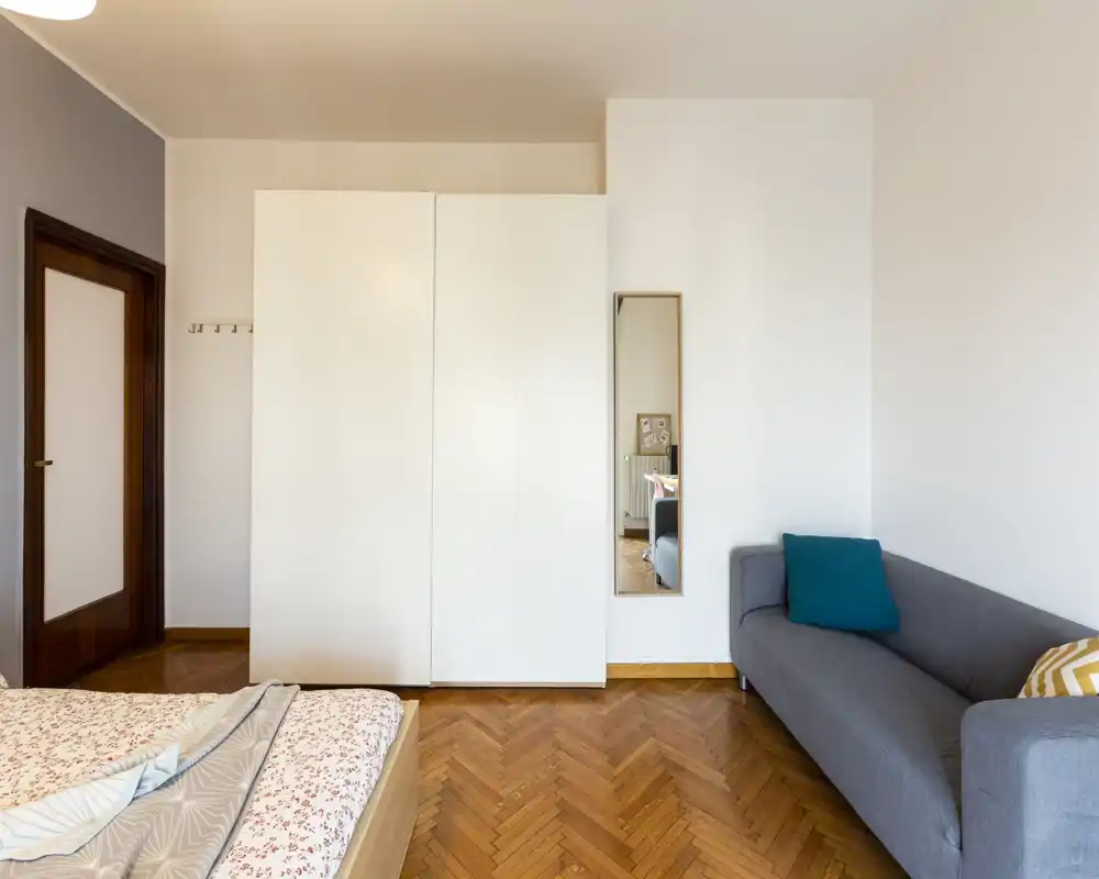 Camera da letto luminosa e arredata con semplicità: letto matrimoniale visibile, grande armadio bianco e divano grigio. Pavimento in parquet a spina di pesce, pareti neutre e tessili creano un ambiente accogliente e ben illuminato.