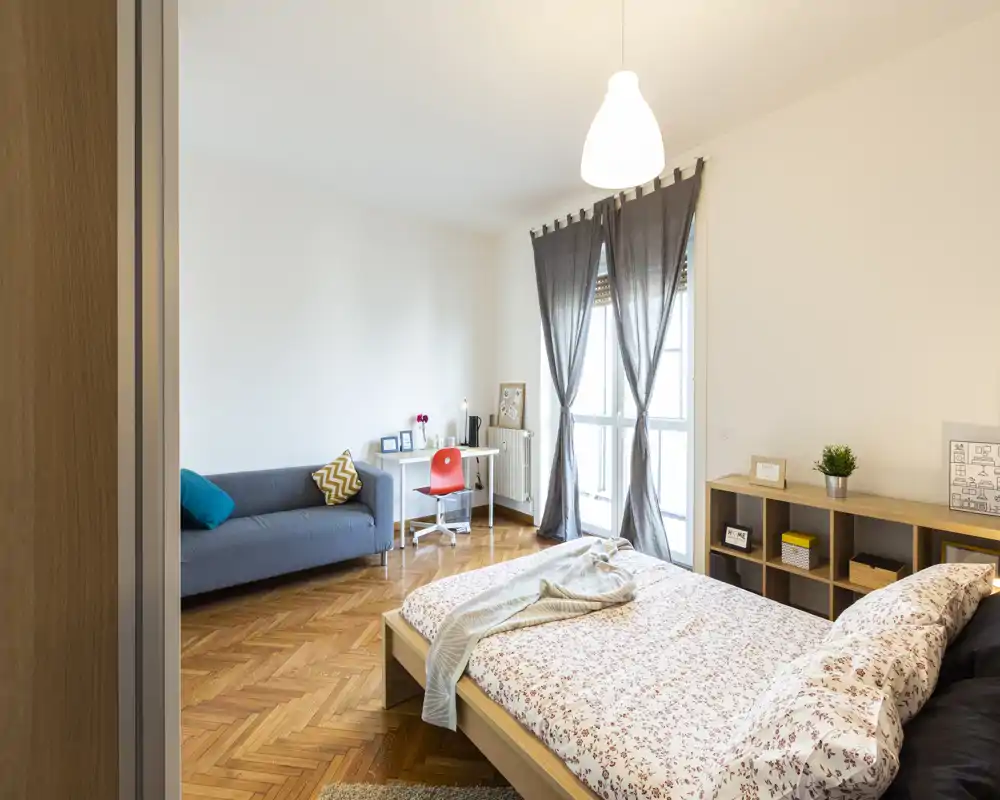 Ampia camera da letto ben illuminata con letto basso in legno, biancheria a fantasia e una coperta accogliente. Grande finestra con tende, una scrivania e un divano creano un'area living/studio versatile.