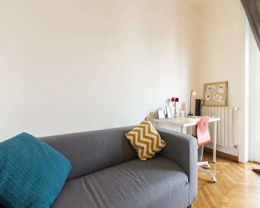 Zona giorno luminosa e moderna con un comodo divano grigio, cuscini colorati e una piccola postazione di lavoro vicino alla finestra — perfetta per rilassarsi o lavorare da casa.