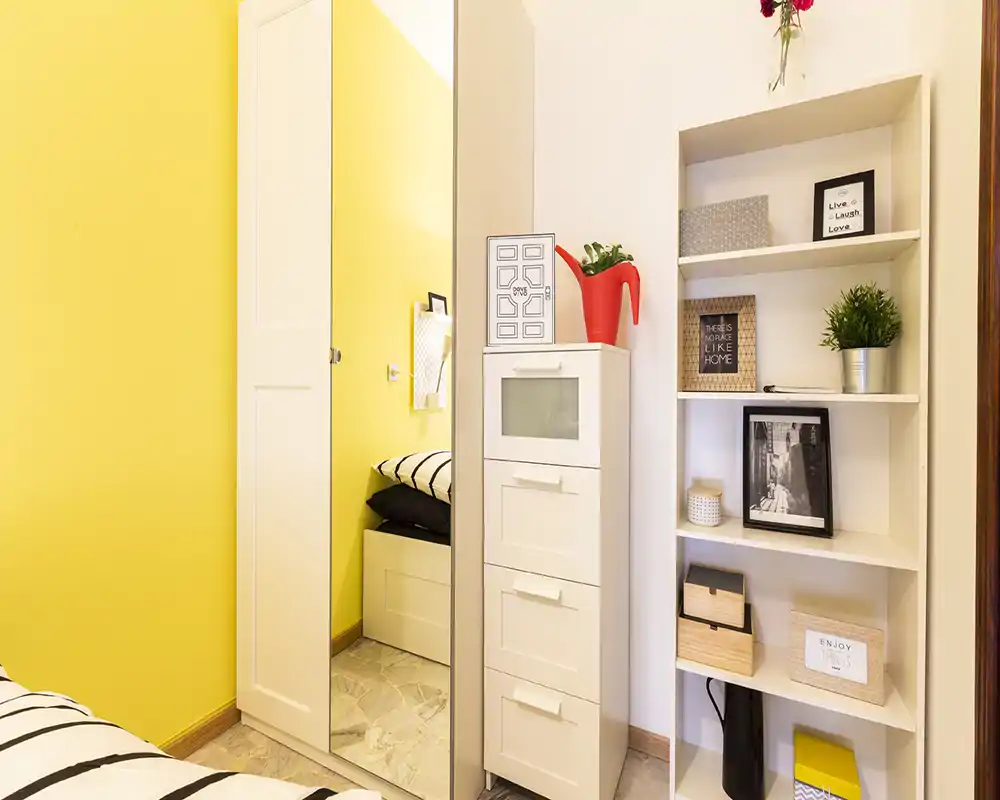 Bright, modern bedroom corner with a mirrored wardrobe, white storage units and decorative shelving; cheerful yellow accent wall and striped bedding add a fresh, cozy feel.