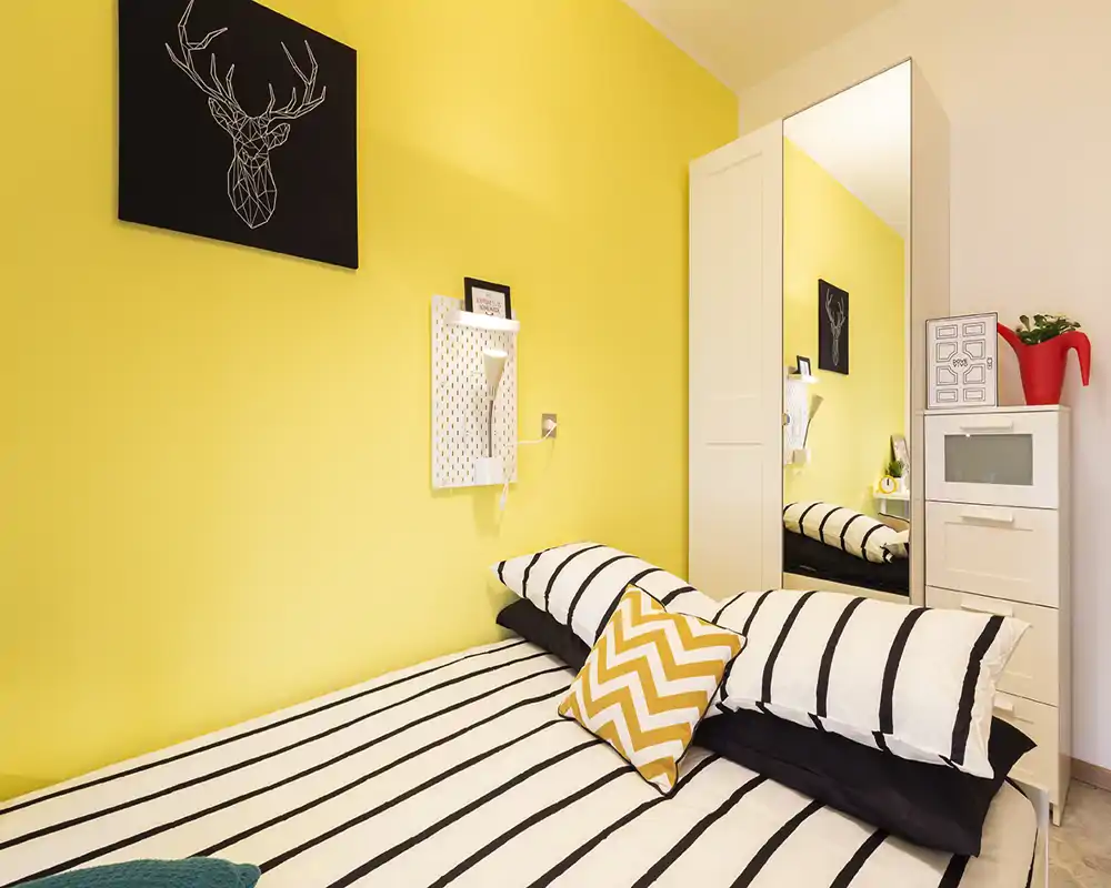 Bright, modern bedroom with a bold yellow accent wall, striped bedding and a chevron cushion. A mirrored wardrobe and compact storage maximize space in this cozy, well-lit sleeping area.