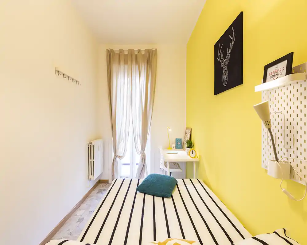 Sunny, modern bedroom with a striped bed in the foreground, bright yellow accent wall and a small workspace by the window.