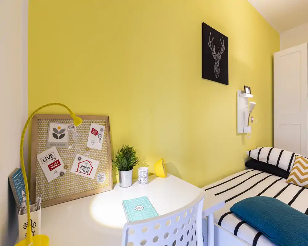 Sunny, well-staged small bedroom corner with a striped single bed, cozy pillows and a white desk with decorative accents — ideal for a student or single occupant.