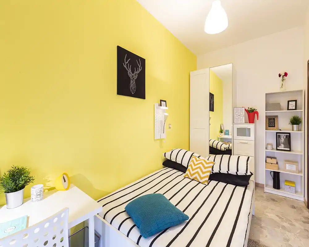 Bright, styled single bedroom with a striped bed, decorative cushions and a cheerful yellow accent wall; includes desk and shelving for storage—ideal for a cozy rental.
