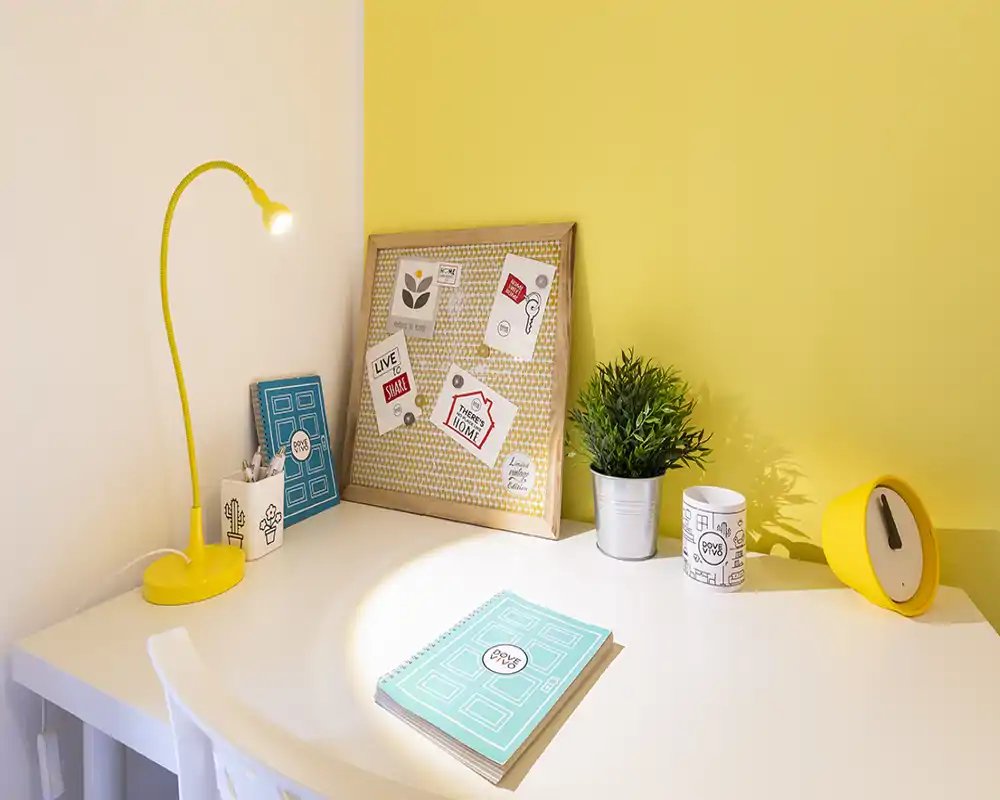 A bright, cheerful desk corner with yellow accents, a small lamp, potted plant and decorative pinboard — ideal for studying or working.