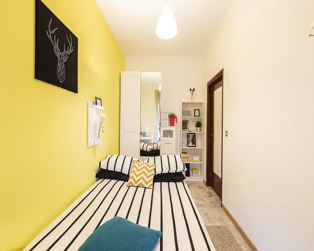 Bright, neatly staged bedroom with a striped double bed against a cheerful yellow accent wall, mirrored wardrobe and compact shelving for storage.
