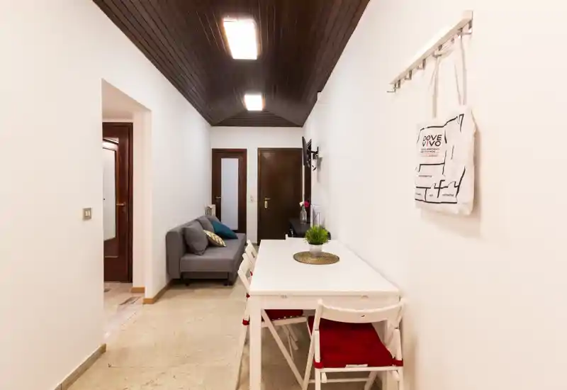 Compact living/dining corridor with a small grey sofa and a white dining table with red-cushioned chairs; bright, clean interior with wooden ceiling accents.