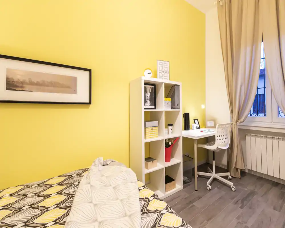 Bright, cozy bedroom with sunny yellow walls, a patterned bedspread and a small work corner with desk and shelving by the window — ideal for a comfortable stay.