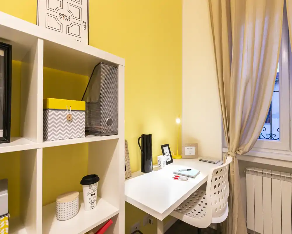 Bright and tidy study corner with a white desk, perforated chair and shelving against a warm yellow wall — ideal for a compact home office.