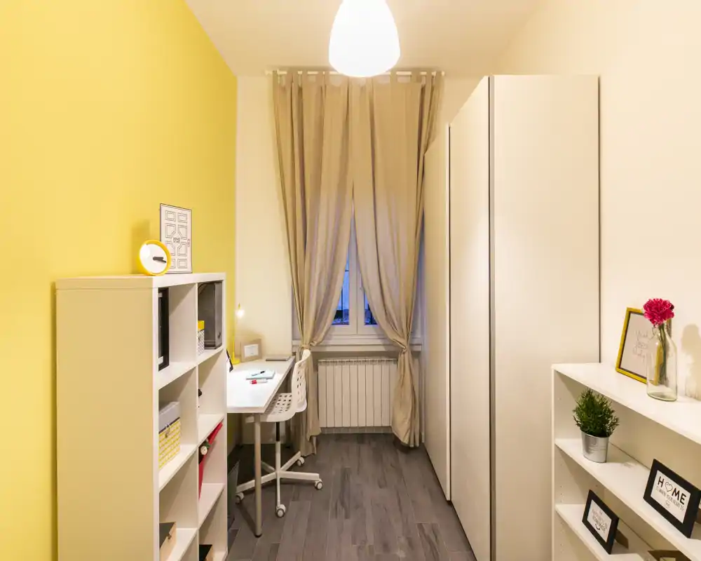 Compact furnished bedroom/home office with warm yellow accent wall, desk by the window, wardrobe and shelving—bright, neatly staged and ideal for a single occupant.